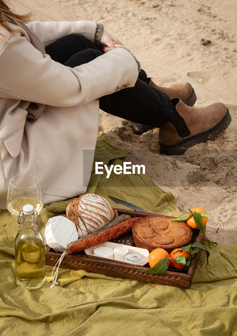 Unrecognizable female in elegant coat sitting on blanket near bottle and glass of white wine and tray with pie with tangerines and bread with cheese and sausages while resting on beach on weekend day