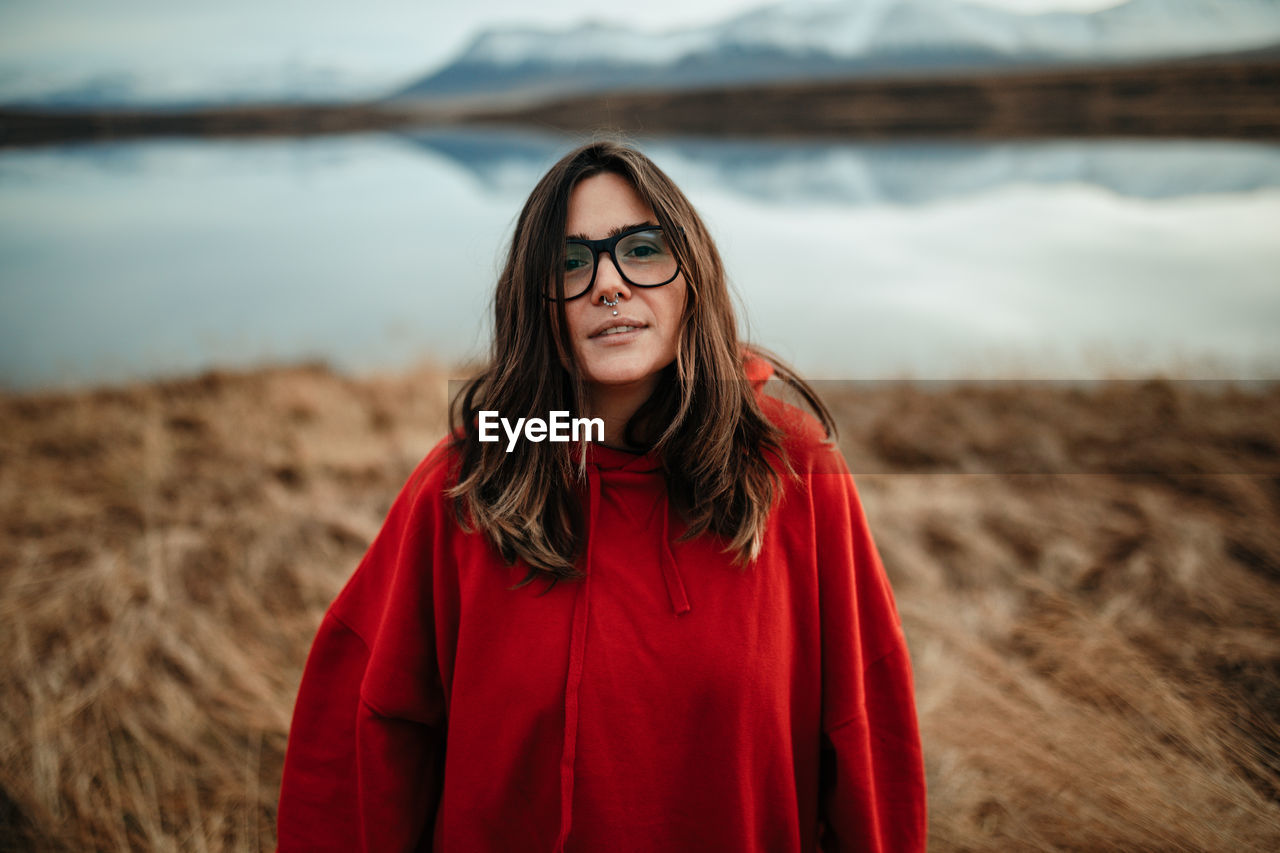 Young laughing tourist in eyeglasses with piercing and windy hair near water in sunny day on blurred background