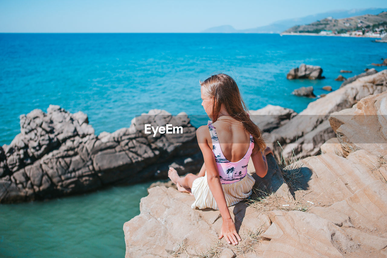 REAR VIEW OF WOMAN SITTING ON ROCK AT SEA SHORE