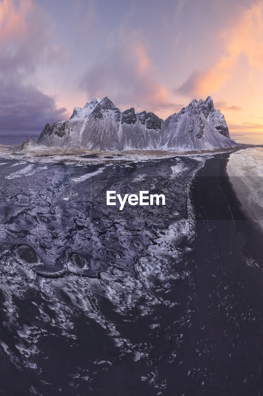 Drone view of calm frozen endless sea surrounding high rocky snowy vestrahorn mountain during picturesque sunset in stockness beach, iceland