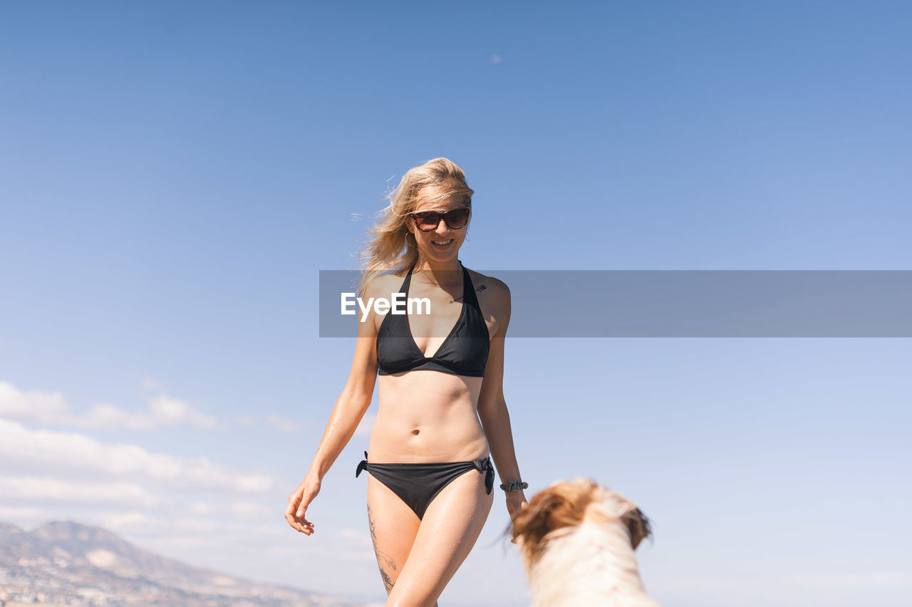 Woman in bikini with dog at beach against blue sky