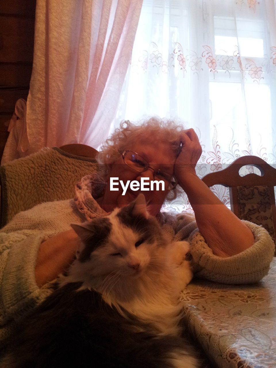 Portrait of smiling senior woman with cat sitting at table against curtain