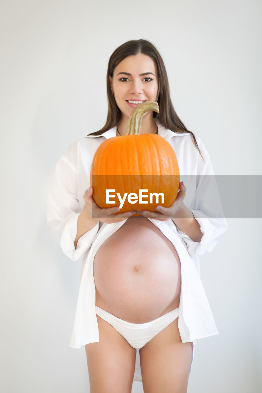 Portrait of smiling pregnant woman holding pumpkin against white background