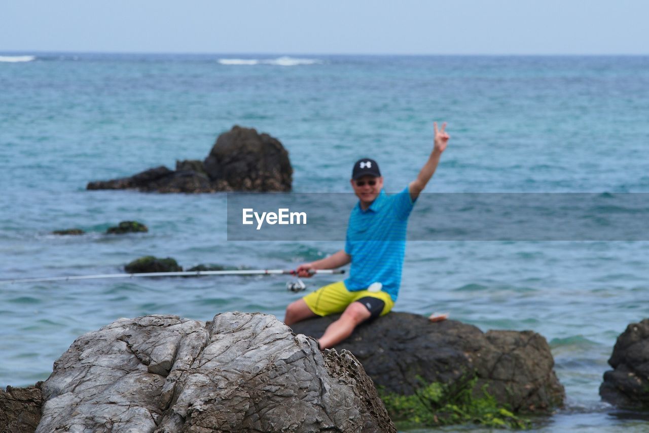 MAN STANDING ON ROCK BY SEA AGAINST CLEAR BLUE SKY