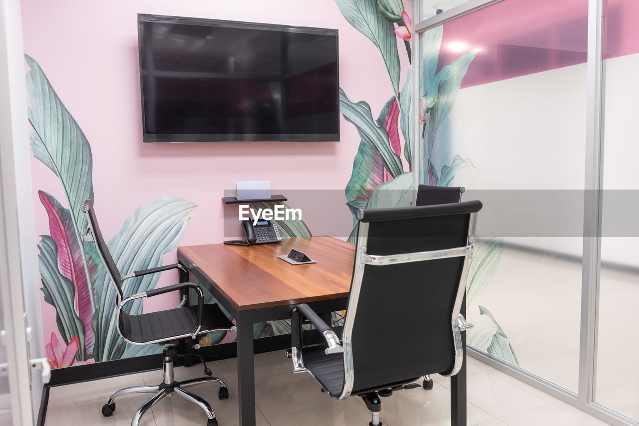 Small wooden conference table with comfortable chairs in creative office with modern tv hanging on wall with floral print