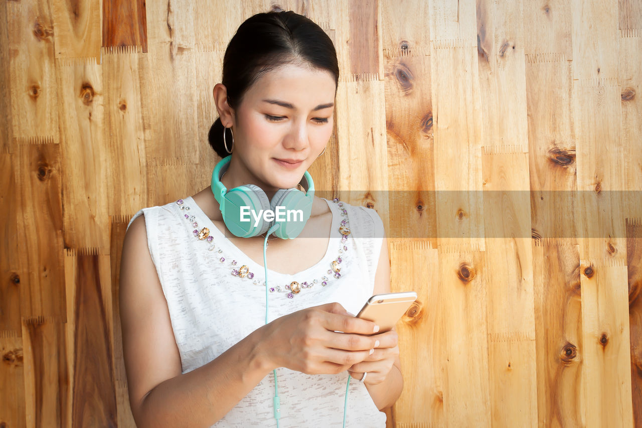 Woman using phone while standing against wooden wall