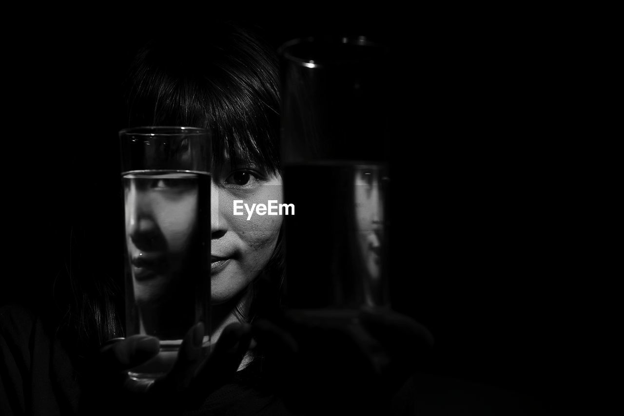 Close-up portrait of young woman looking through drinking glass against black background