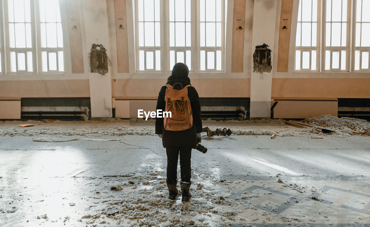 Rear view of young woman with backpack and camera standing in abandoned room