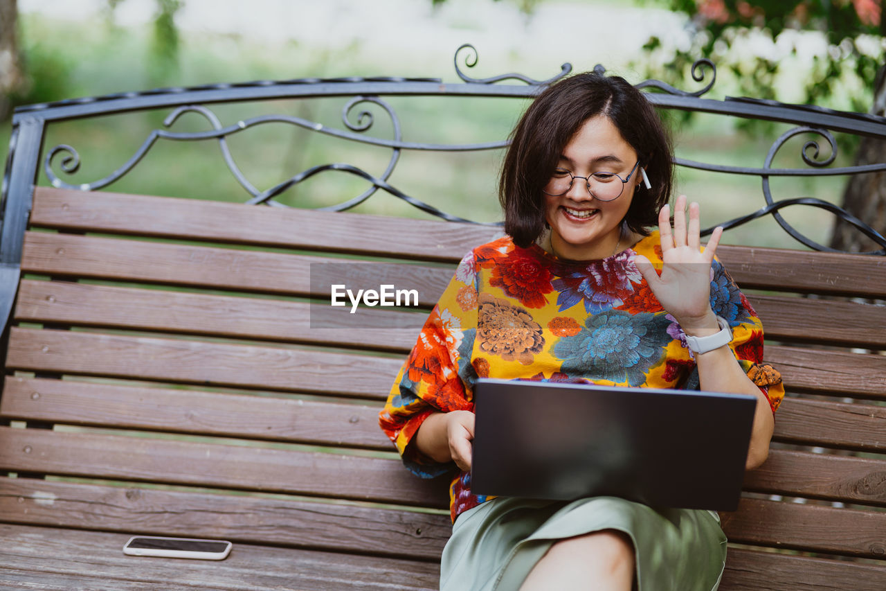 An adult asian woman in a park on bench communicates via video call on a laptop