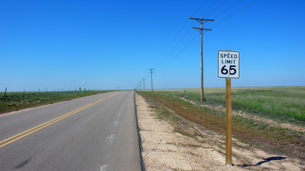 Speed limit 65 sign and electricity pylon by country road against clear blue sky