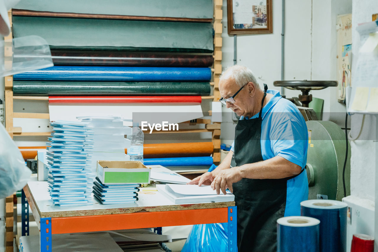 Side view of concentrated senior male artisan with white hair in apron and eyeglasses standing at table with pile of papers and notebook in printing workshop