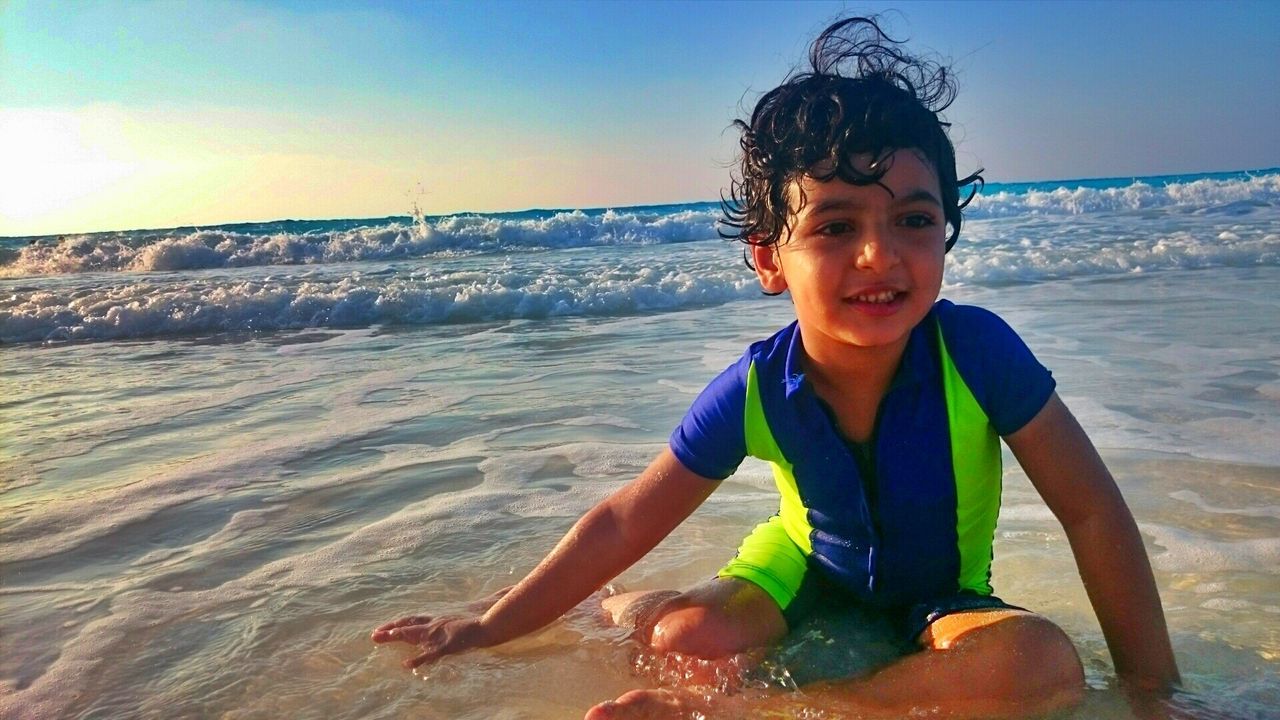 Cute boy sitting on shore at beach against sky during sunset