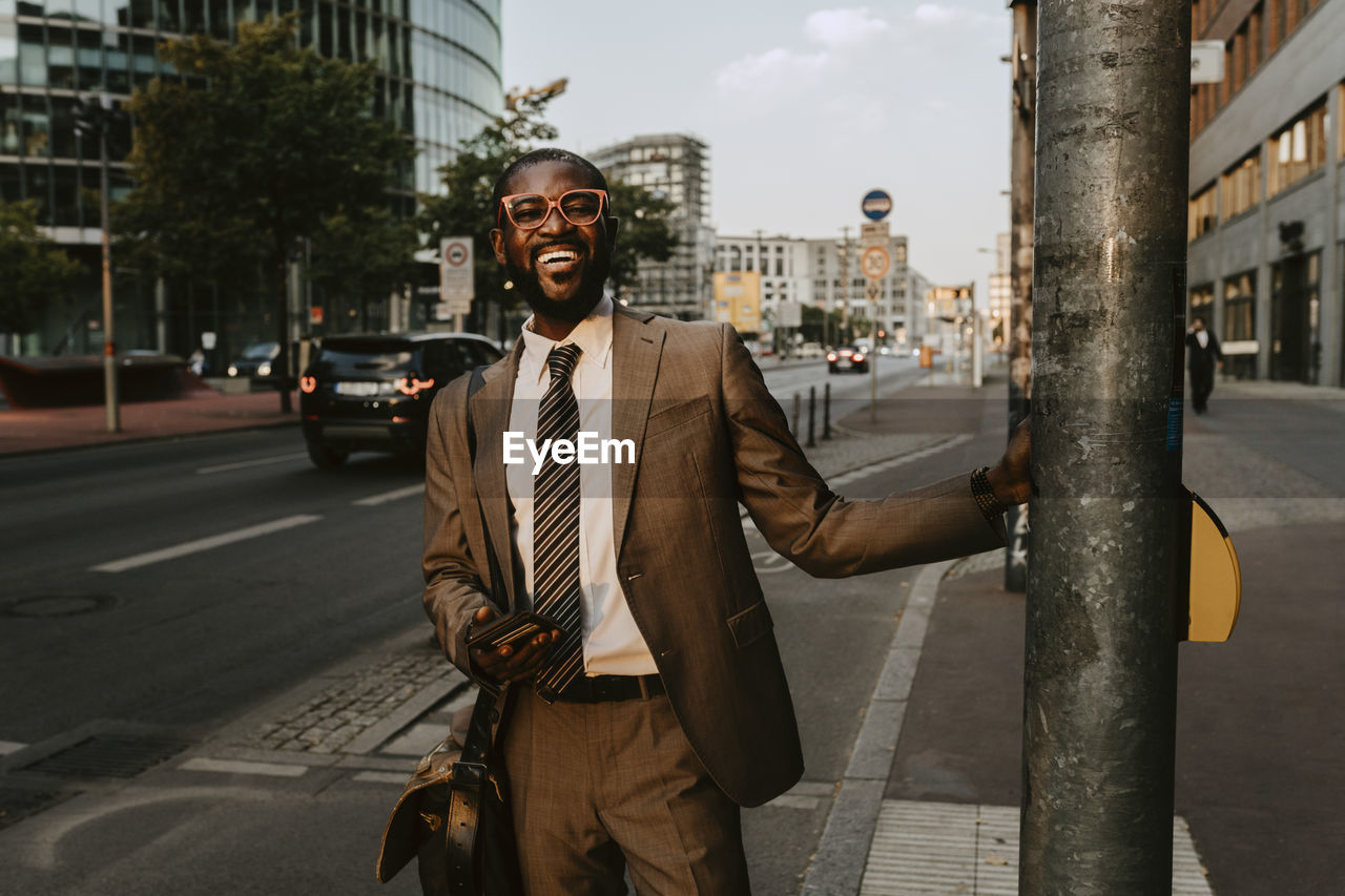 Smiling male entrepreneur standing by pole on footpath in city