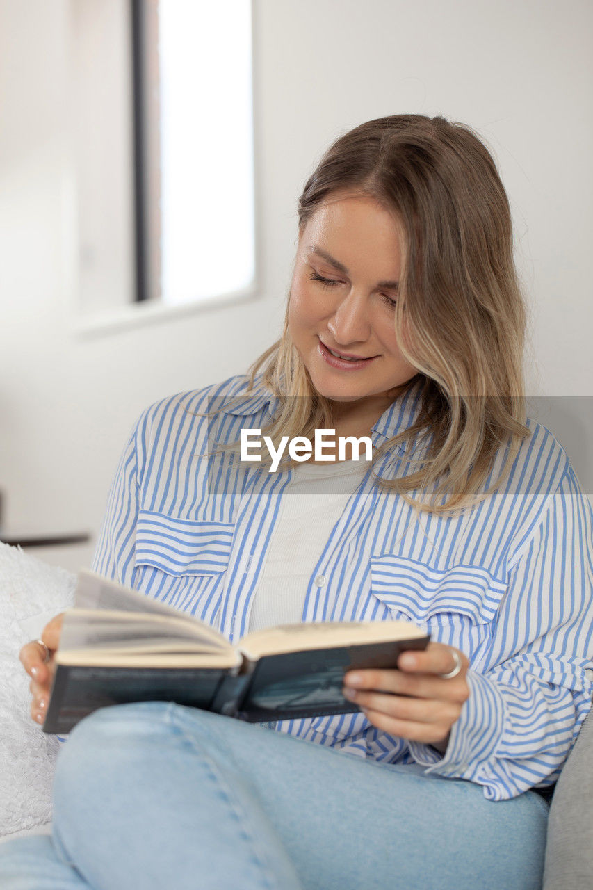sitting, one person, adult, women, indoors, casual clothing, reading, smiling, domestic life, communication, furniture, technology, holding, looking, domestic room, lifestyles, person, sofa, living room, happiness, home interior, wireless technology, relaxation, three quarter length, clothing, looking down, female, emotion, long hair, blond hair, young adult, hairstyle, front view, striped, enjoyment, publication, mobile phone, digital tablet, convenience, internet, activity, leisure activity, cheerful, portable information device, smartphone, comfortable, portability, business, book