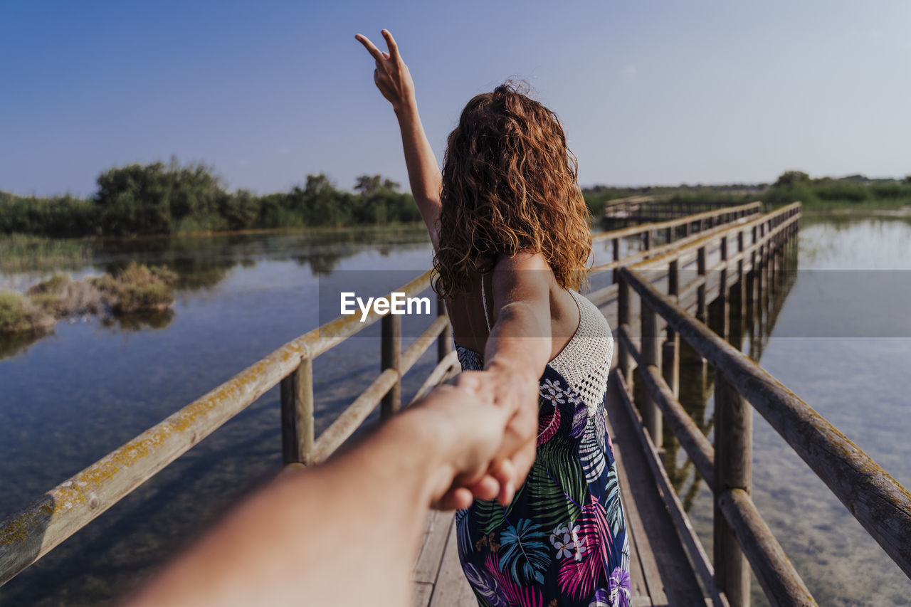 Woman holding hand of boyfriend while standing on footbridge against sky