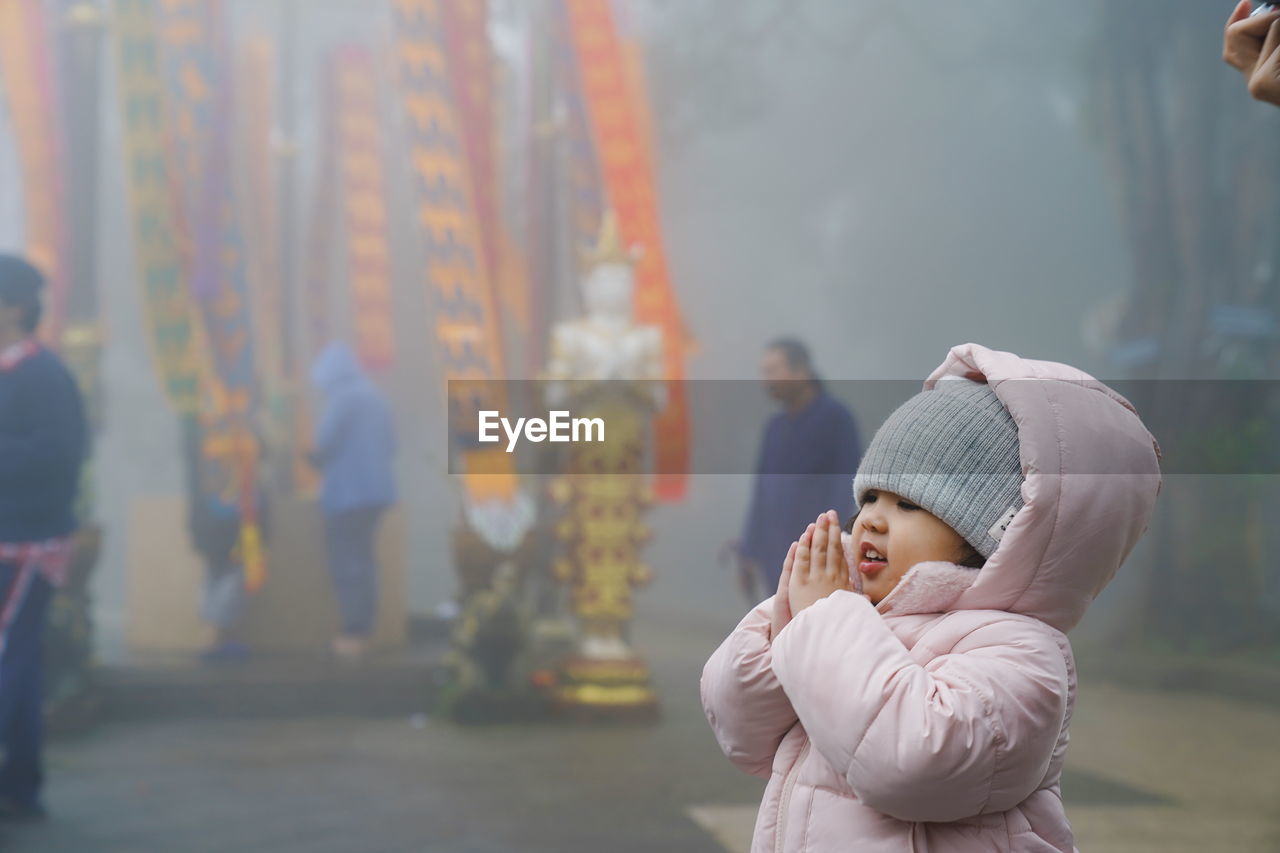 Cute girl wearing warm clothing praying while standing during foggy weather