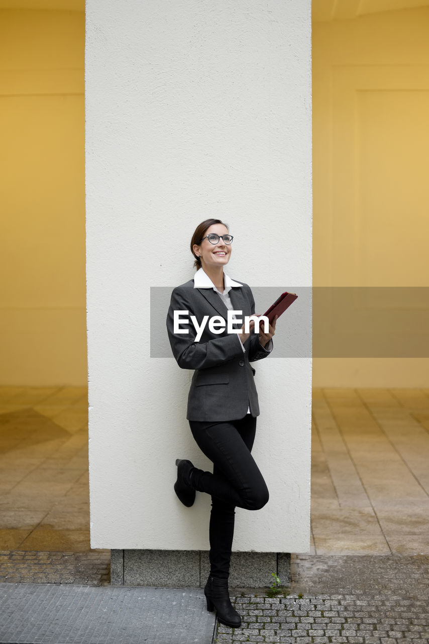 Full length of smiling businesswoman holding digital tablet while standing against wall