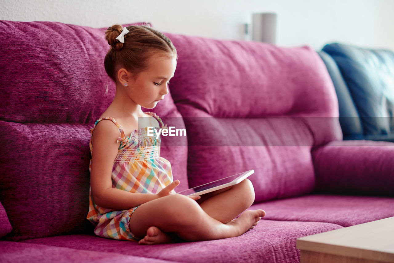Cute girl using digital tablet sitting on pink sofa at home