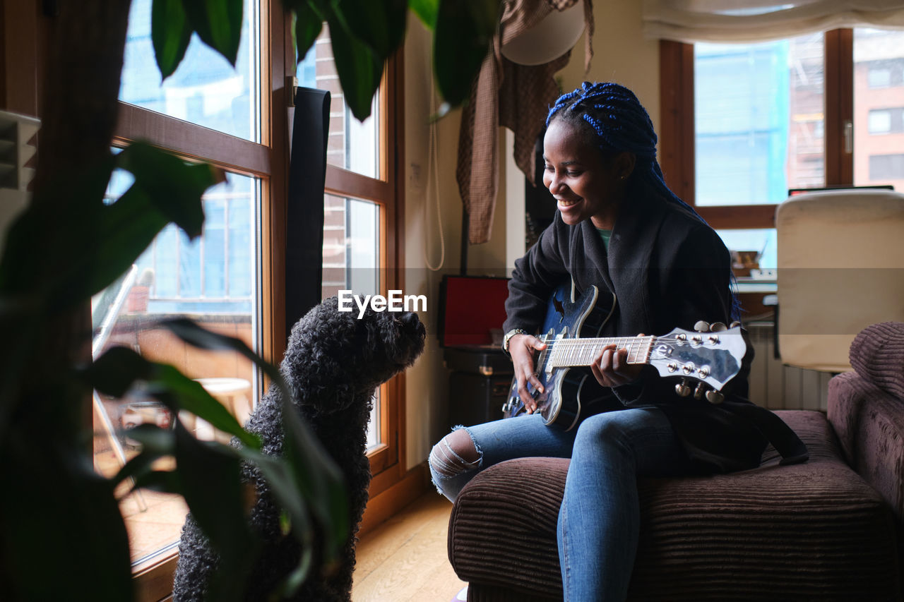 Smiling african american young female musician with dreadlocks wearing jacket and jeans playing guitar while sitting on sofa opposite poodle dog in living room