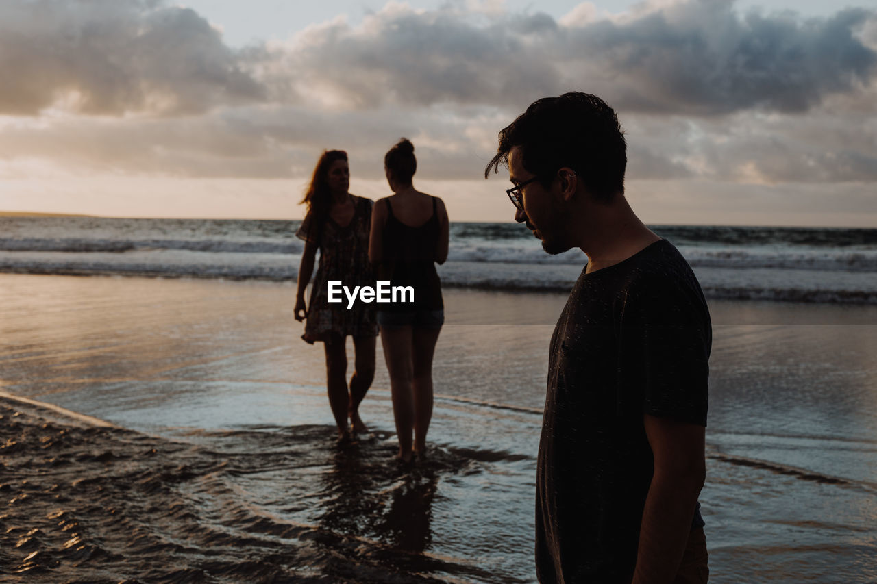 Friends standing on beach against sky during sunset