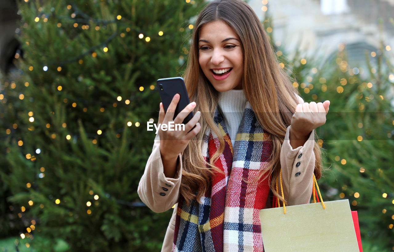 Smiling woman using mobile phone while standing against christmas tree