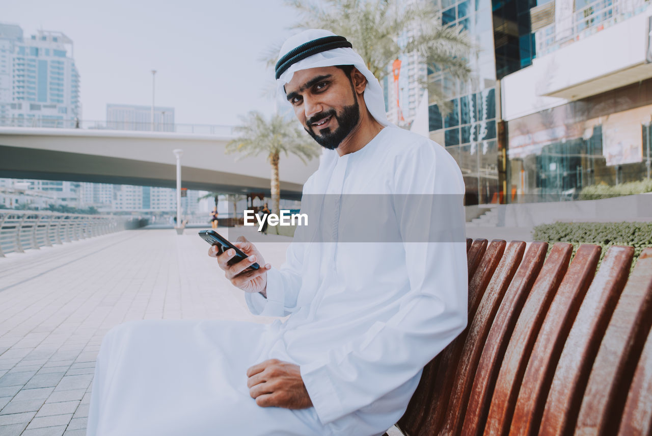 Young man using smart phone sitting on bench in city