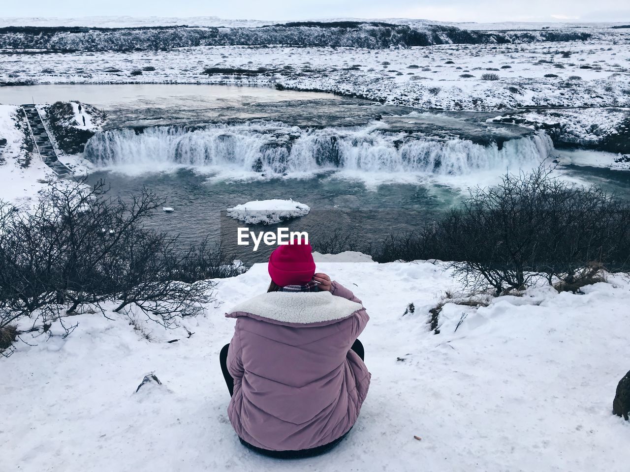 Rear view of person looking at waterfall while sitting on snow covered field