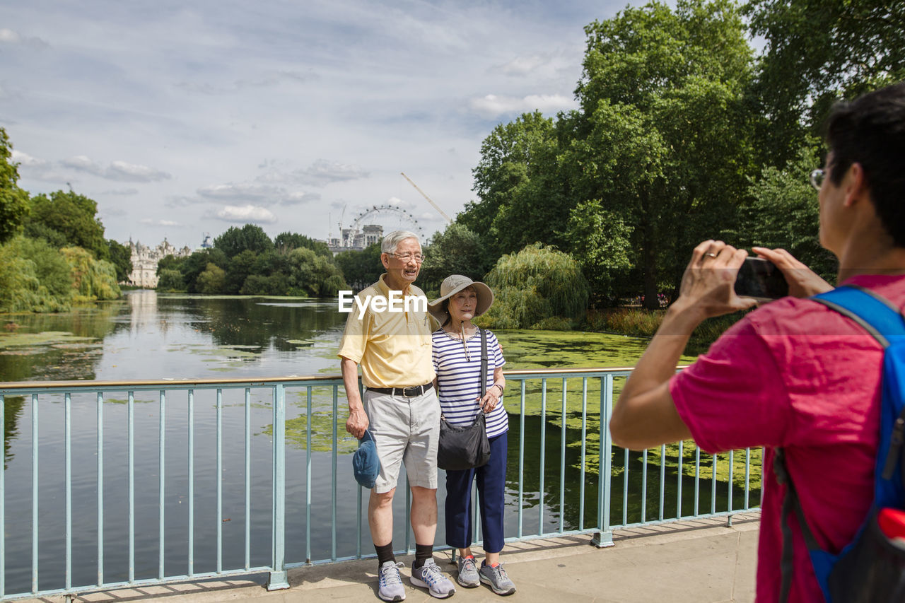 Son photographing parents with mobile phone standing on footbridge over river against sky at park during sunny day