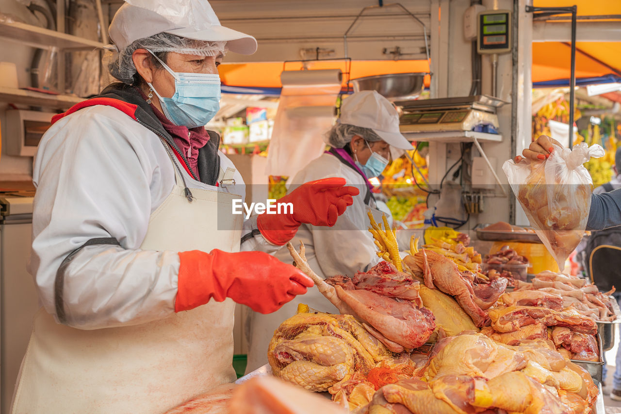 Peruvian female sellers in face masks and aprons standing behind counter with raw chicken meat during work on itinerant fair