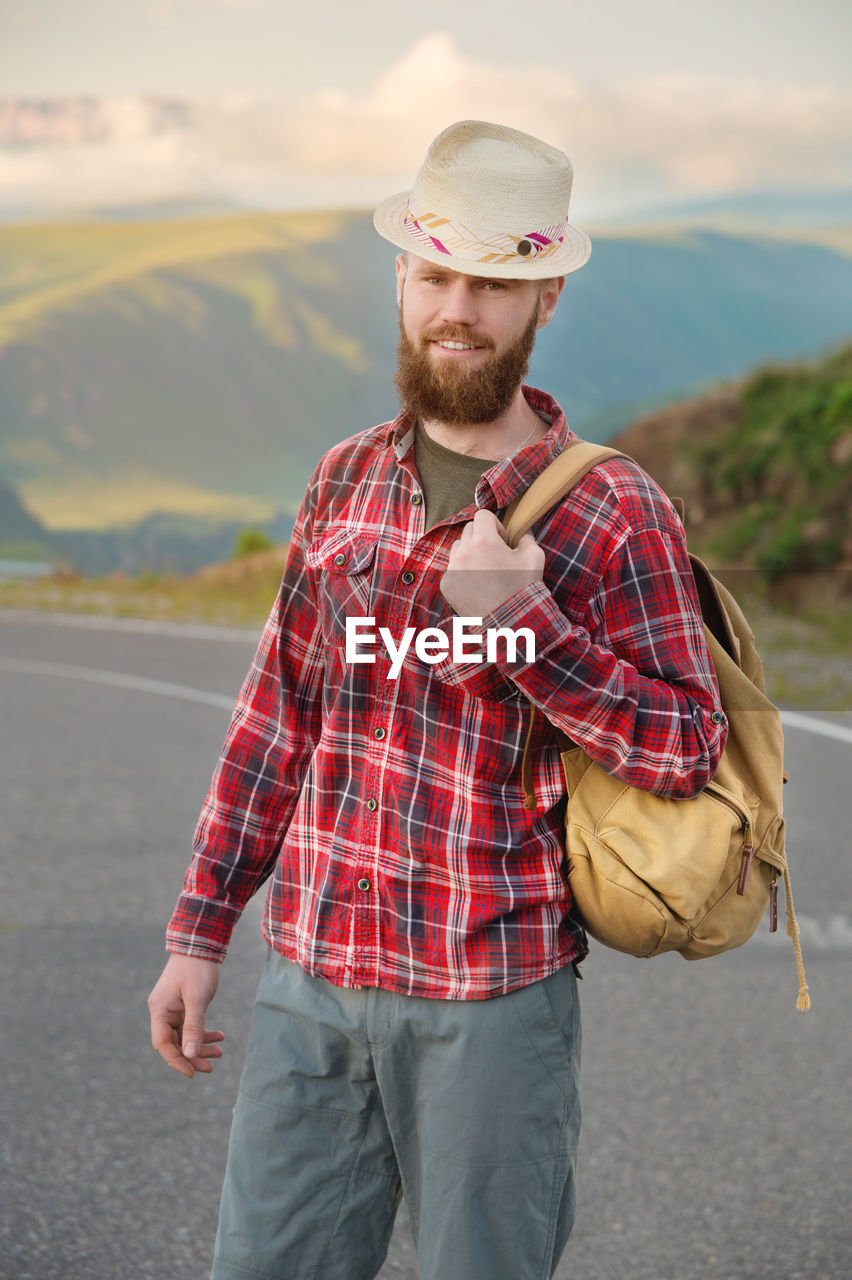 Portrait of a sly cheerful bearded jewish man hitchhiker in a hat and shirt with a backpack against 