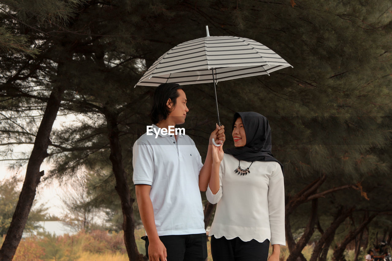 Young couple holding umbrella while standing against trees