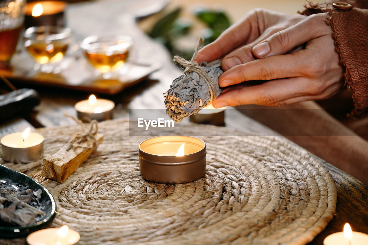 Woman hands burning white sage, before ritual on the table with candles and green plants. smoke of