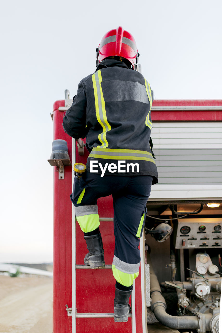 Back view of anonymous firefighter wearing red protective helmet and uniform standing on fire truck ladder and looking into the distance in daytime