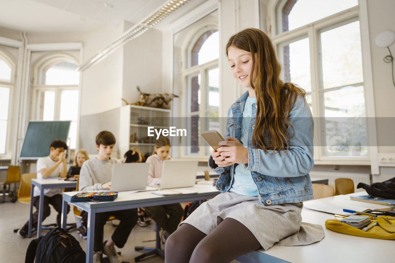 Smiling female student using smart phone while sitting at desk in classroom