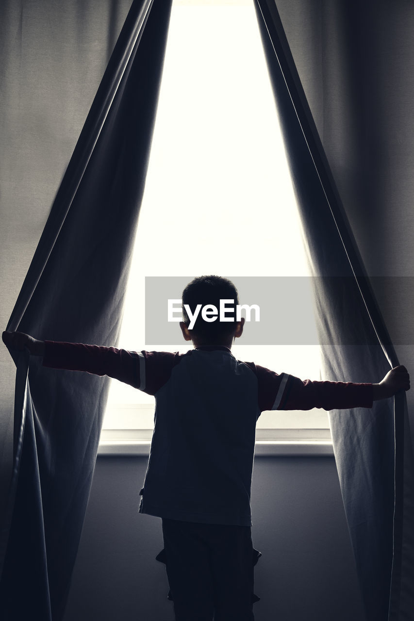 Rear view of boy holding curtains while looking through window at home