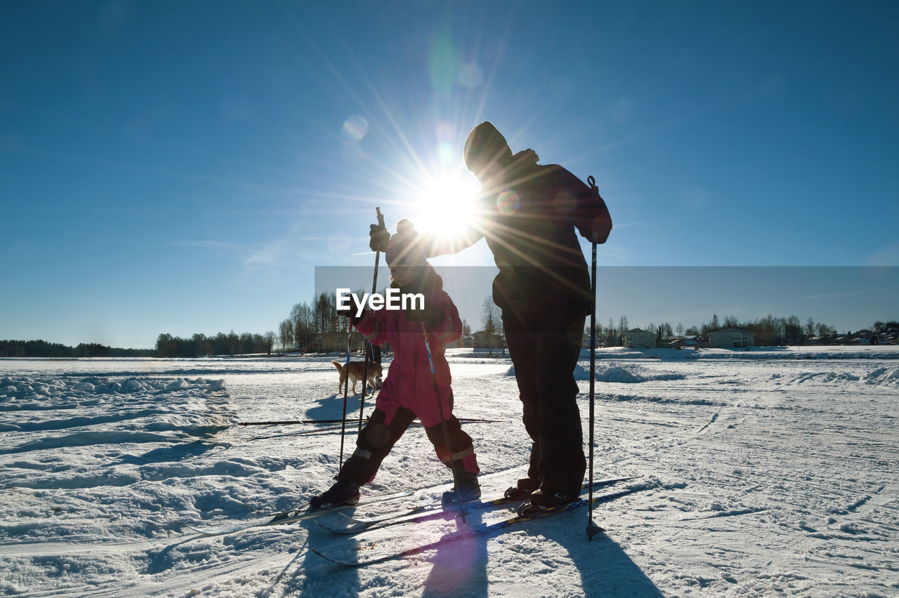 Mother assisting daughter during ski against bright sky