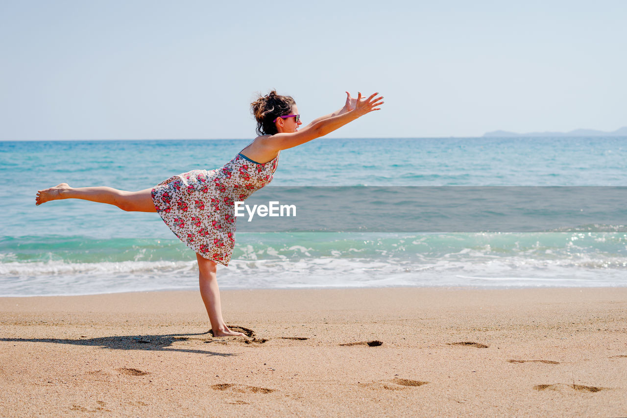 Woman with arms raised exercising while standing on one leg at beach against sky