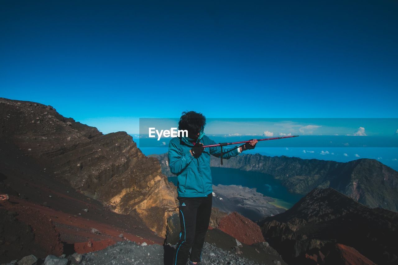 Young man aiming with hiking pole on mountain against blue sky