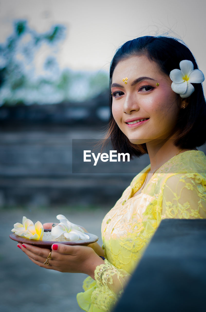 The woman wearing traditional chlothing from bali with carrying offerings is looking to the camera