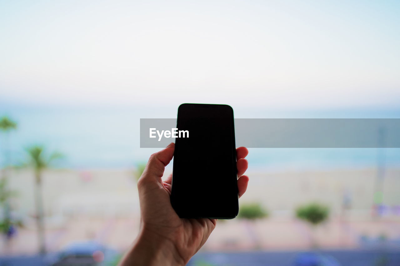 Close-up of mans hand photographing sea against sky