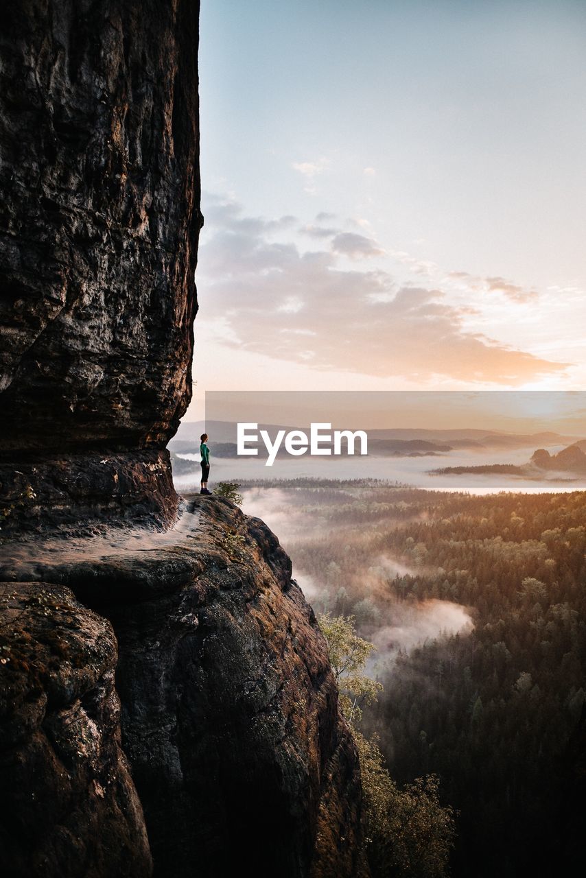 Side view of woman standing on rock against sky during sunset