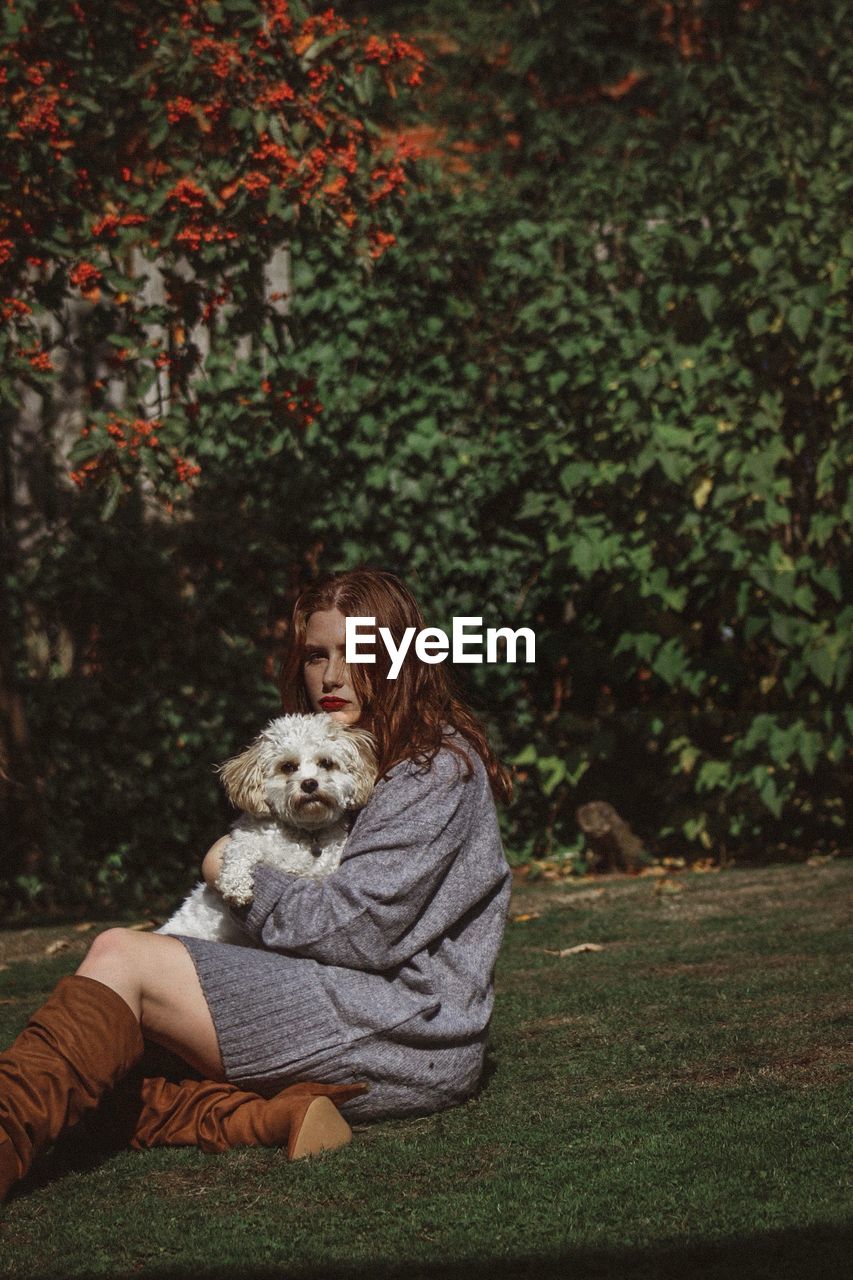 Portrait of young woman holding dog while sitting on grassy field against trees