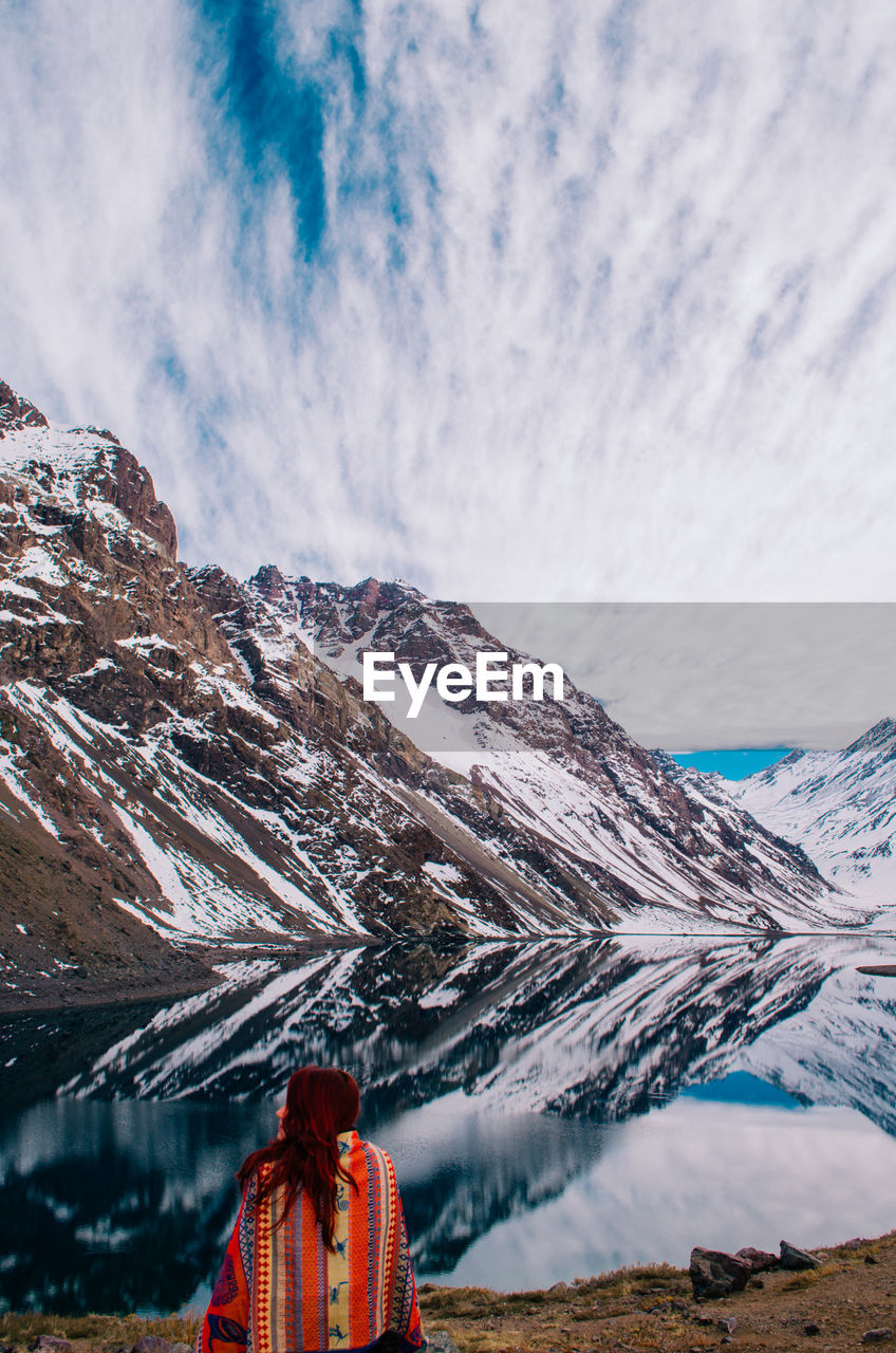 Rear view of woman sitting by lake against mountain and sky