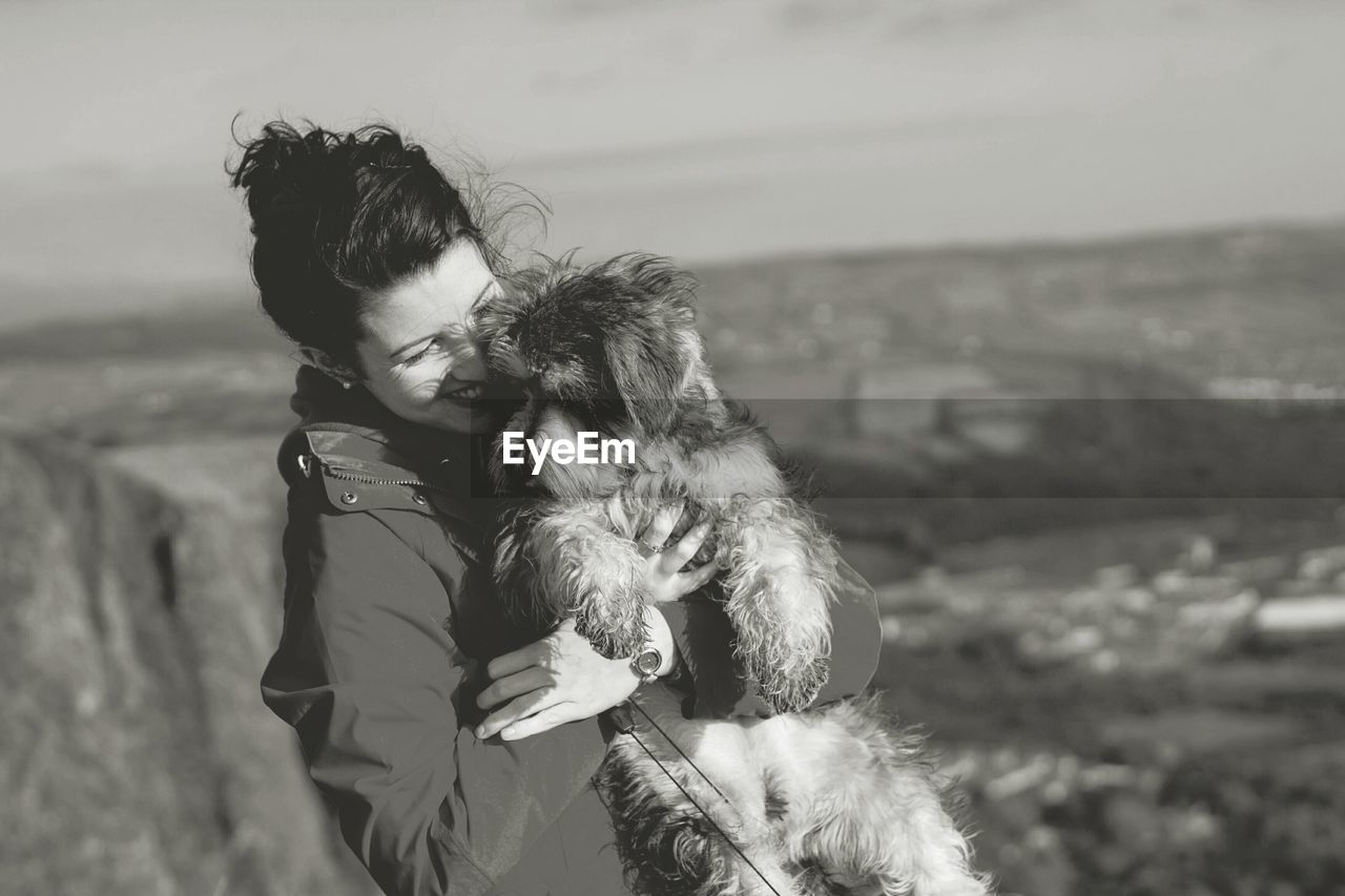 Happy woman carrying dog on field against sky