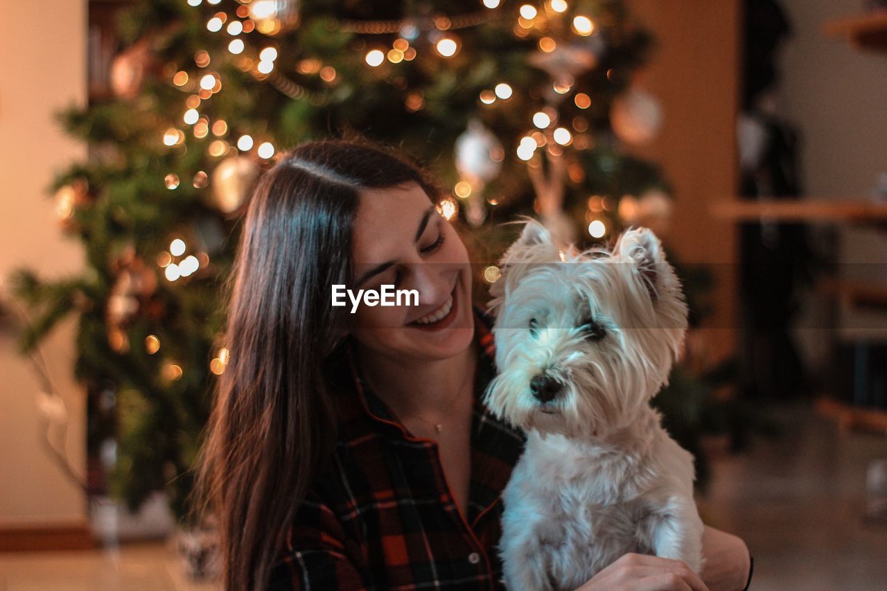 Smiling woman with dog against christmas tree at home