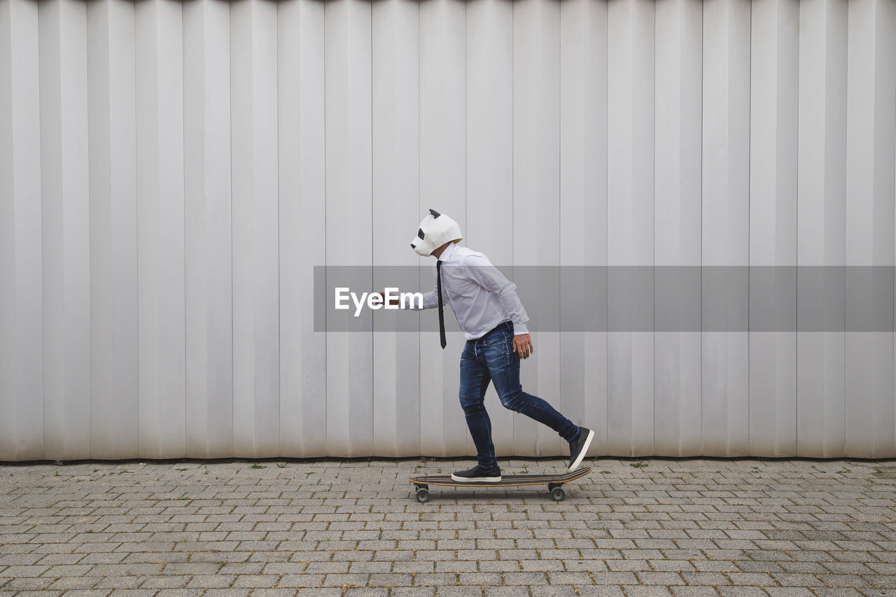 Businessman skateboarding on longboard with a panda bear mask against gray wall person