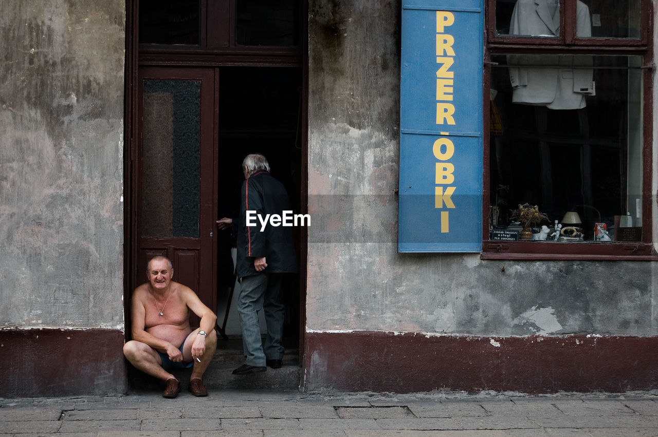 MAN AND WOMAN SITTING ON STREET AGAINST WINDOW