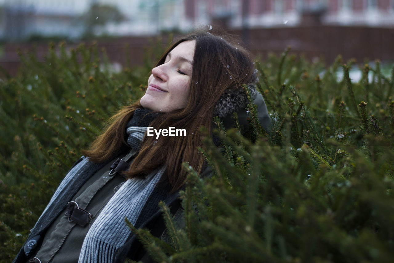 Smiling woman with closed eyes leaning on plants at park during winter