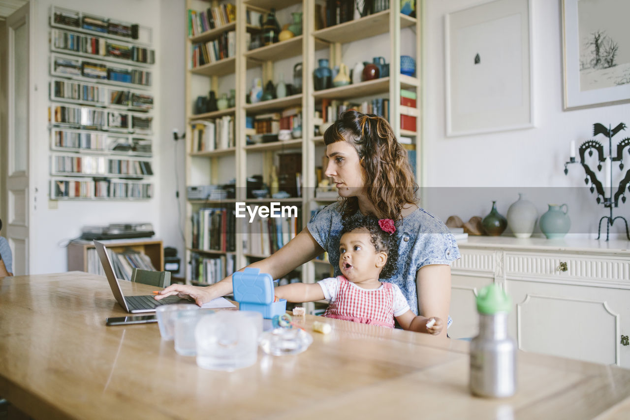 Woman using laptop while sitting with daughter at dining table in house