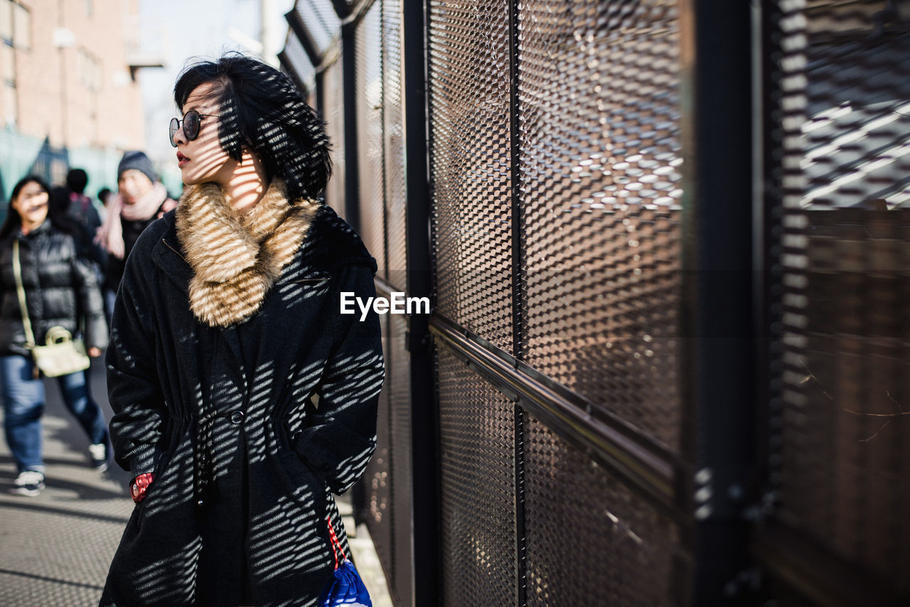 Young woman in warm clothing wearing sunglasses standing by built structure